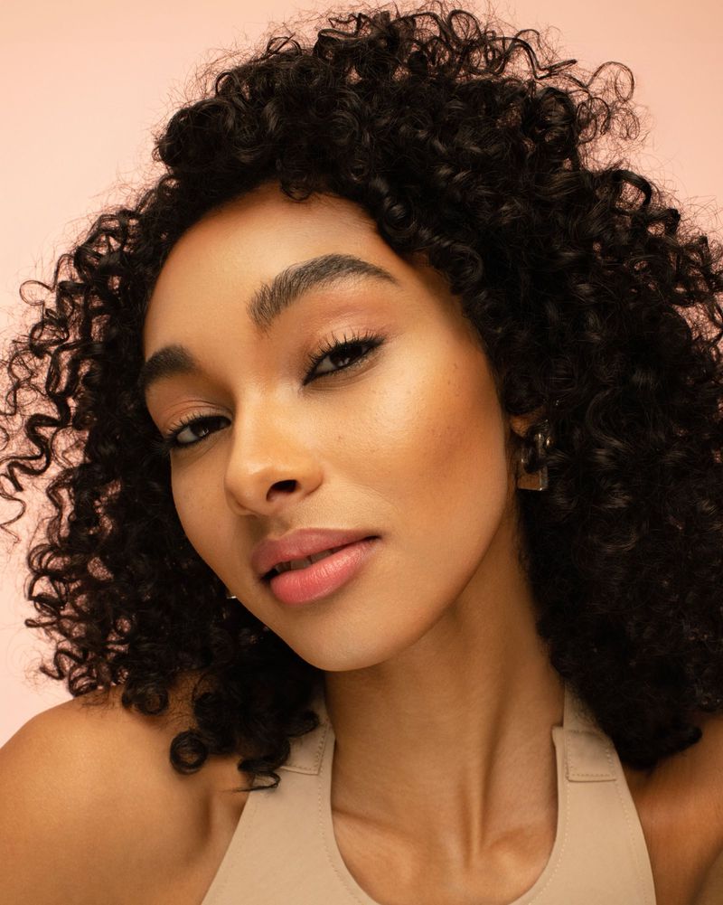 Close-up studio shot of a smiling African-American woman with curly hair, light makeup, and a beige top, set against a light pink background.