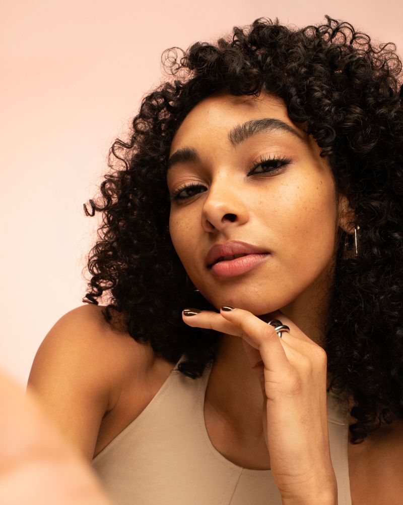 Close-up portrait of a woman with curly hair, wearing a neutral top, with a hand near her chin, against a soft pink background.