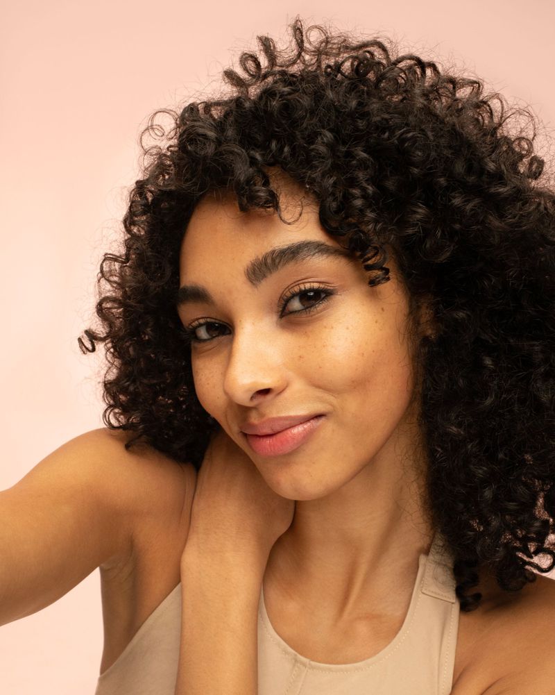 Close-up portrait of a smiling woman with curly hair. Soft, natural lighting and a muted pink backdrop create a serene and inviting atmosphere.