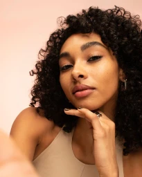 Close-up portrait of a woman with curly black hair resting her chin on her hand, wearing a beige top against a neutral background.