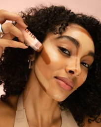 Woman applying contour stick makeup to her cheek with visible cosmetic swatches on her face against a pink studio backdrop.