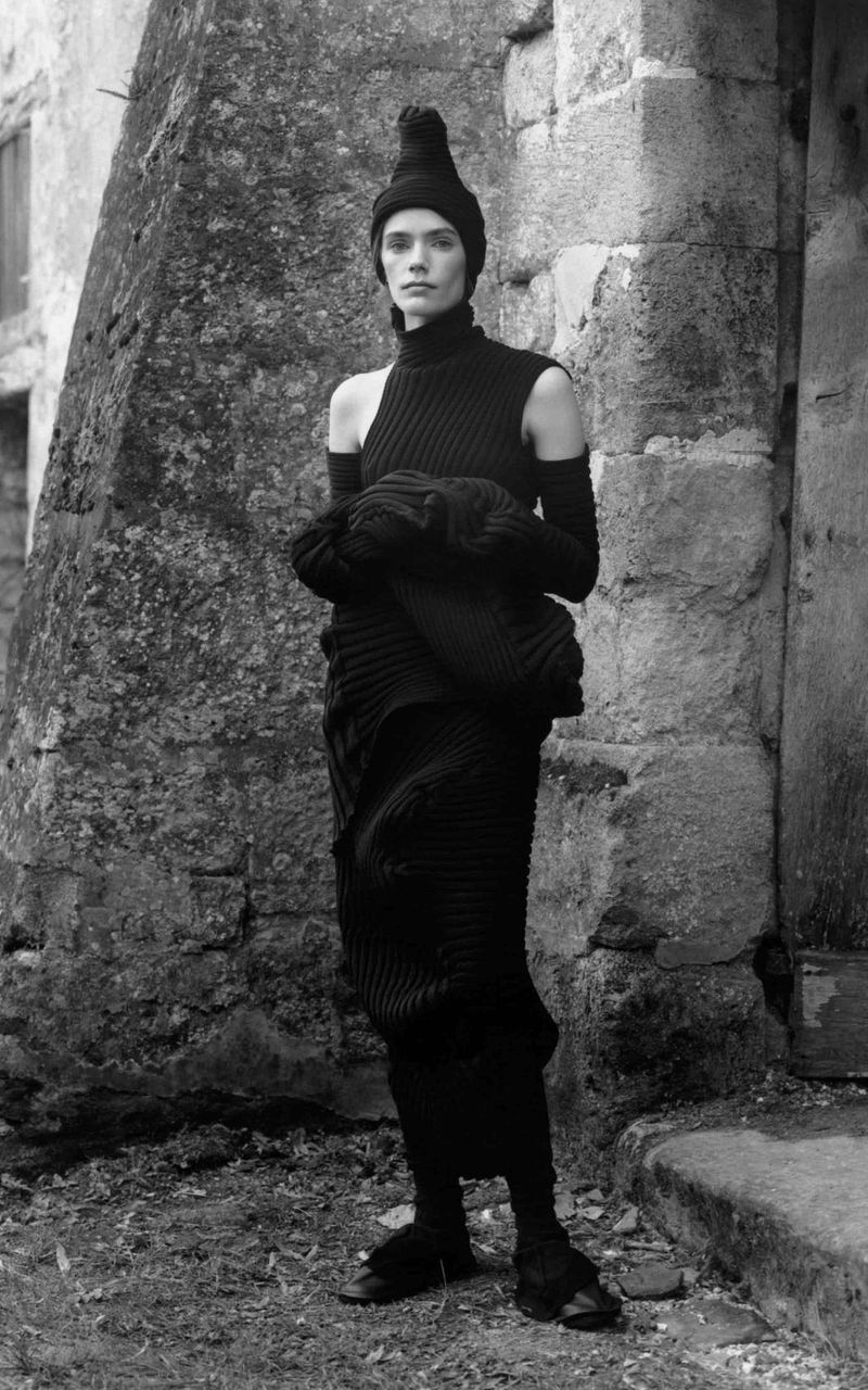 A slender adult female model stands against a weathered stone wall in an outdoor fashion shot featuring an unusual black knit outfit and headdress.