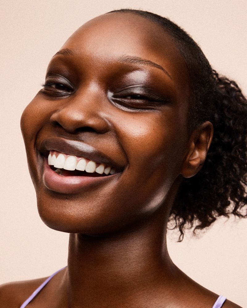 Close-up shot of a smiling adult female with dark skin and curly hair, wearing a purple top against a neutral background.