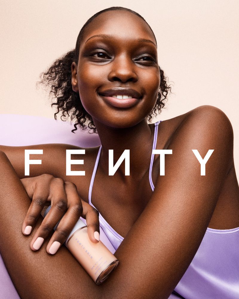 Close-up of a smiling African-American woman wearing a lavender top and holding a foundation bottle, with the 'FENTY' logo in white.