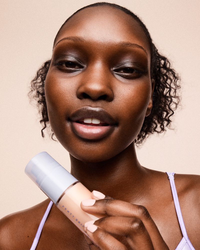 Close-up of an adult woman with dark skin holding a bottle of Fenty Skin in front of a light-beige indoor background, smiling slightly.