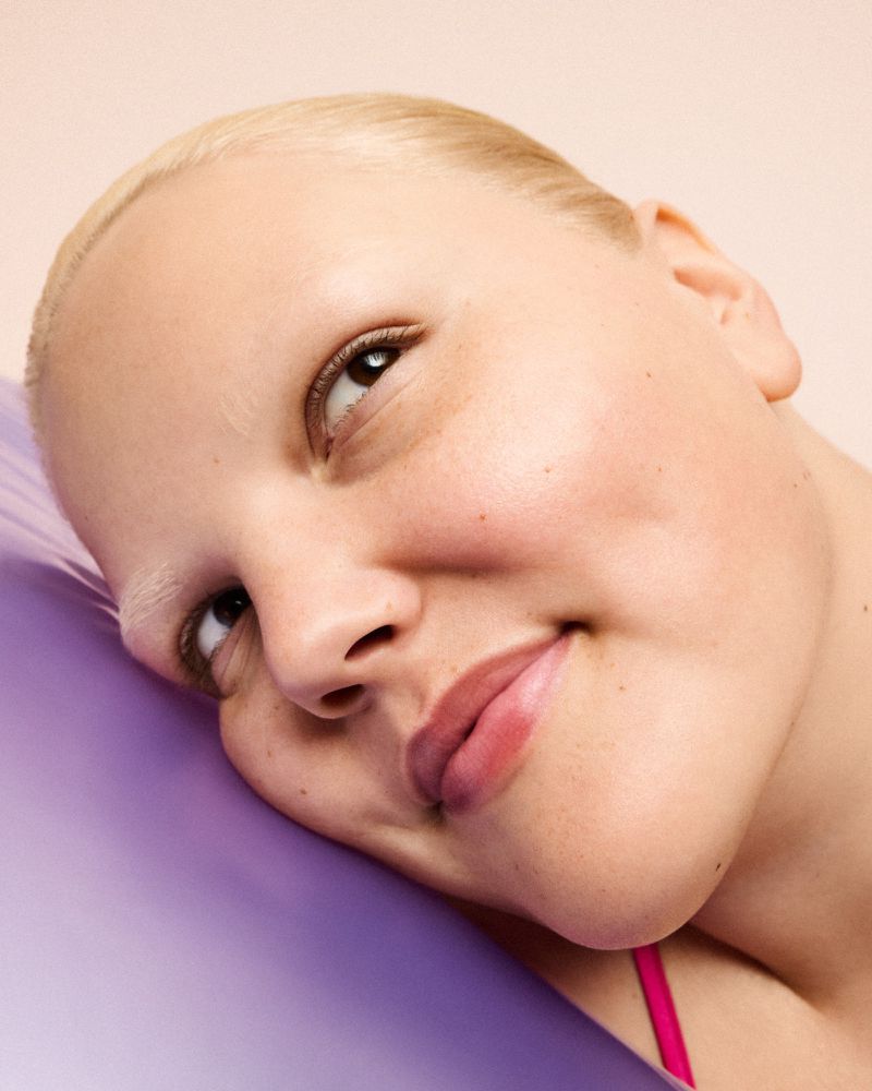 Close-up portrait of a smiling adult woman with short blonde hair, lying on a purple cushion in a brightly lit indoor setting.