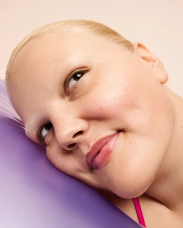 Close-up portrait of a smiling adult woman with short blonde hair, lying on a purple cushion in a brightly lit indoor setting.