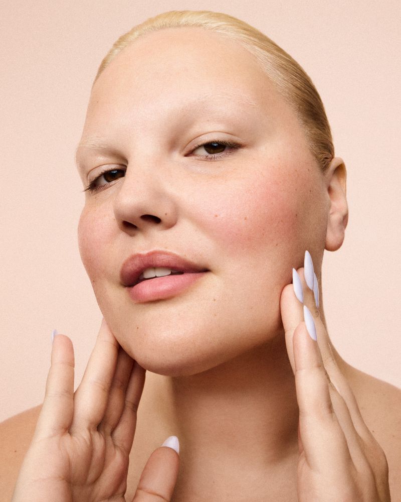 Close-up studio shot of a woman with short blonde hair touching her face with painted nails against a peach backdrop. Beauty shot.