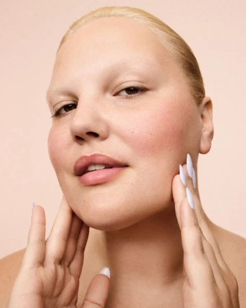 Close-up studio shot of a woman with short blonde hair touching her face with painted nails against a peach backdrop. Beauty shot.