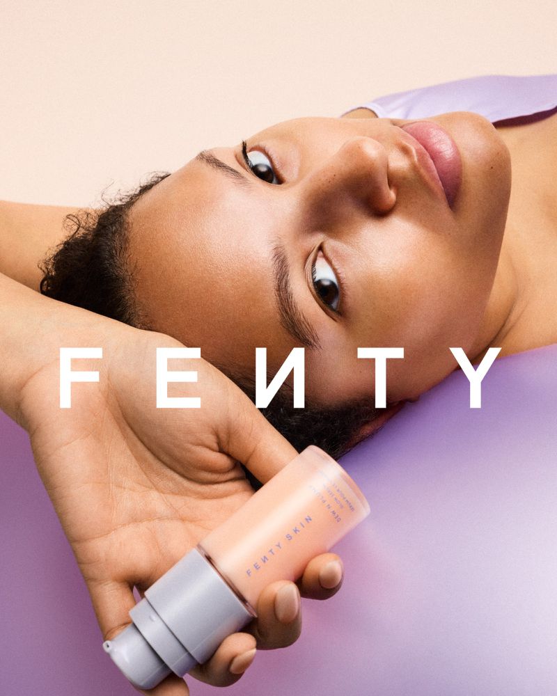 An adult female with short curly hair is angled in a beauty product ad, holding a serum by Fenty Skin against a neutral backdrop.
