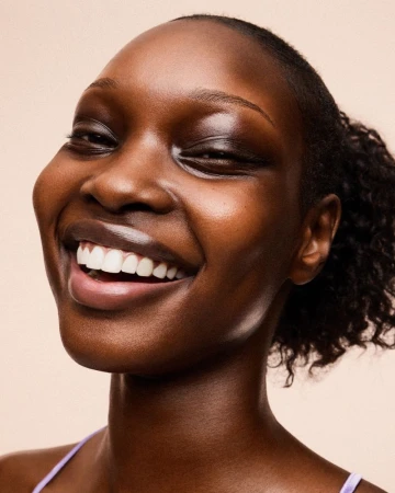 Close-up beauty portrait of a smiling Black woman with dewy, glowing skin against a warm beige seamless background.