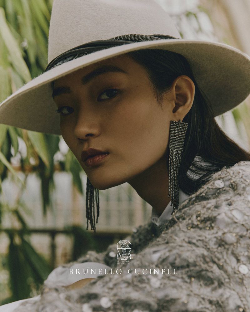 Close-up fashion shot of an Asian woman wearing a wide-brimmed hat and beaded fringe earrings, accessorized with a textured knit sweater.