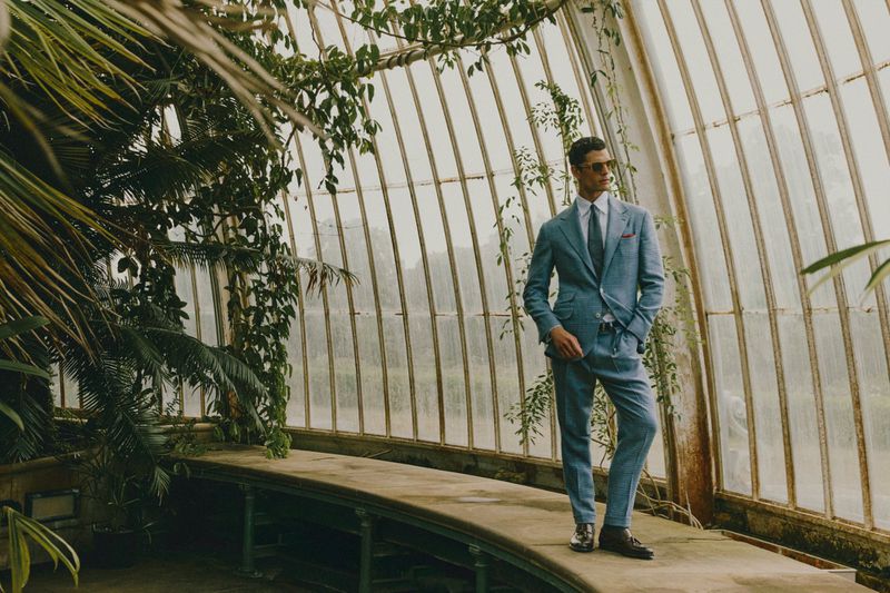 Man in a blue suit posing inside a sunlit greenhouse surrounded by plants, wearing sunglasses and a tie, looking stylish and fashionable.