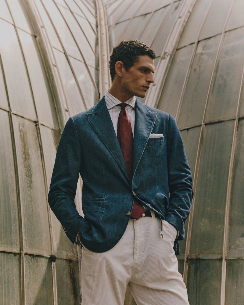 Male model poses outdoors wearing a blue suit jacket, striped dress shirt, red tie, and light beige dress pants in a fashion editorial.