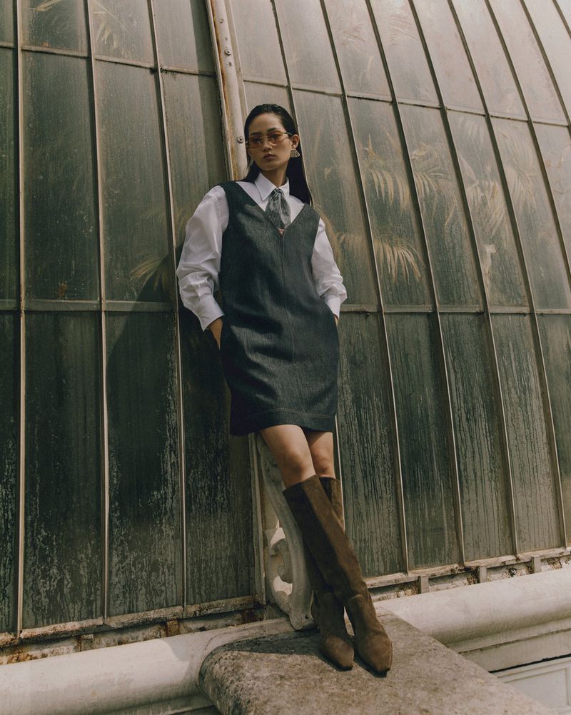 Woman in dress and boots poses against greenhouse windows. Sunlight creates shadows in an outdoor fashion shot.
