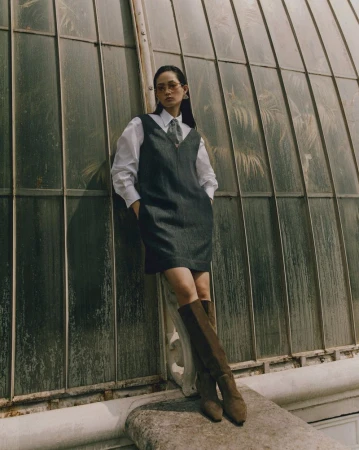 Woman in dress and boots poses against greenhouse windows. Sunlight creates shadows in an outdoor fashion shot.