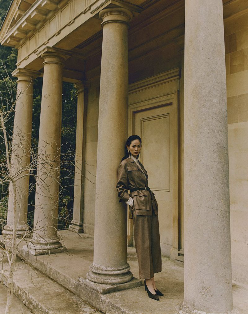 Model Hyunji Shin wearing a brown belted pantsuit leaning against a classical stone column outdoors.