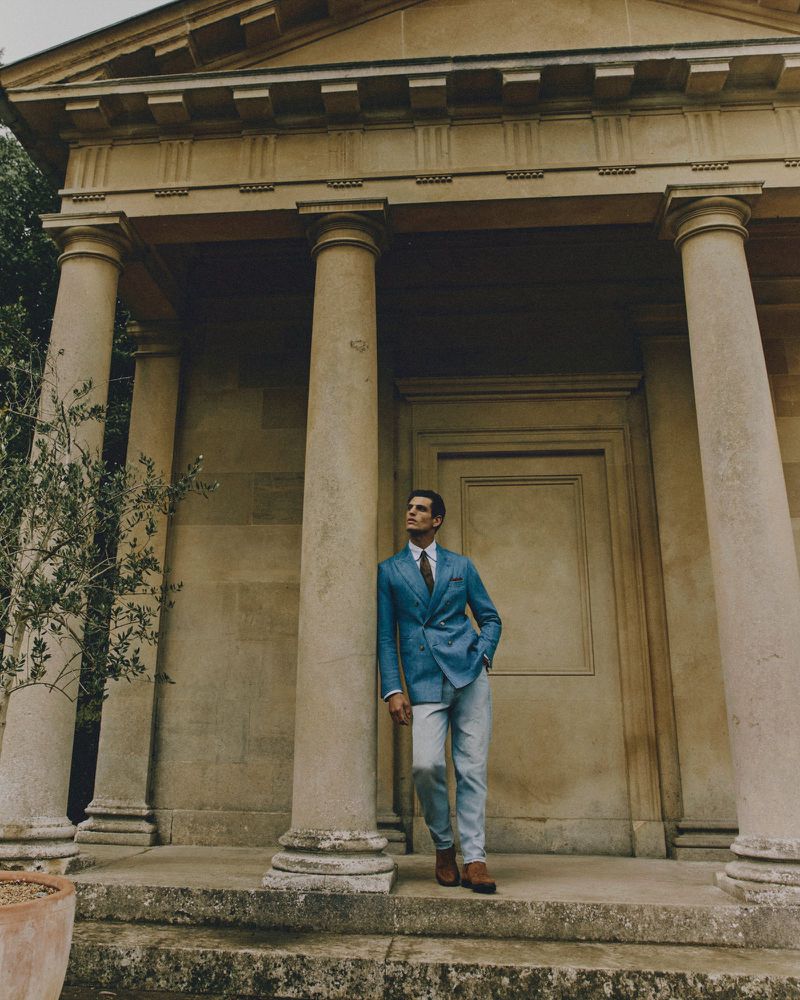 A man in a blue suit stands in front of a stone building with columns, looking to the side, bathed in soft, natural light.