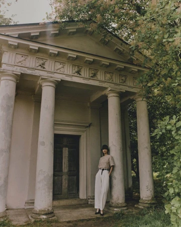 Woman in casual wear poses outdoors next to neoclassical building with columns surrounded by trees, creating an editorial fashion image.