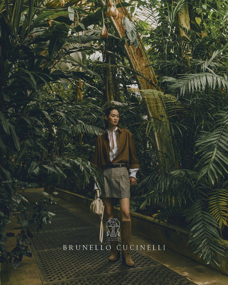 Fashion shot of a woman in a brown shirt, plaid skirt, and boots, carrying a purse in a greenhouse with lush green plants.
