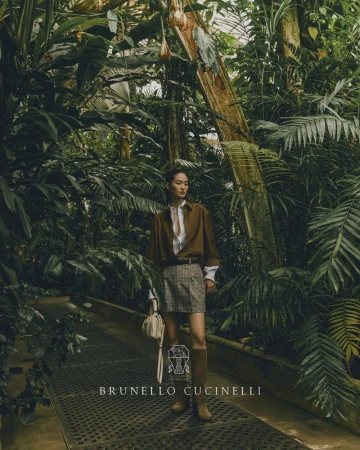 Fashion shot of a woman in a brown shirt, plaid skirt, and boots, carrying a purse in a greenhouse with lush green plants.