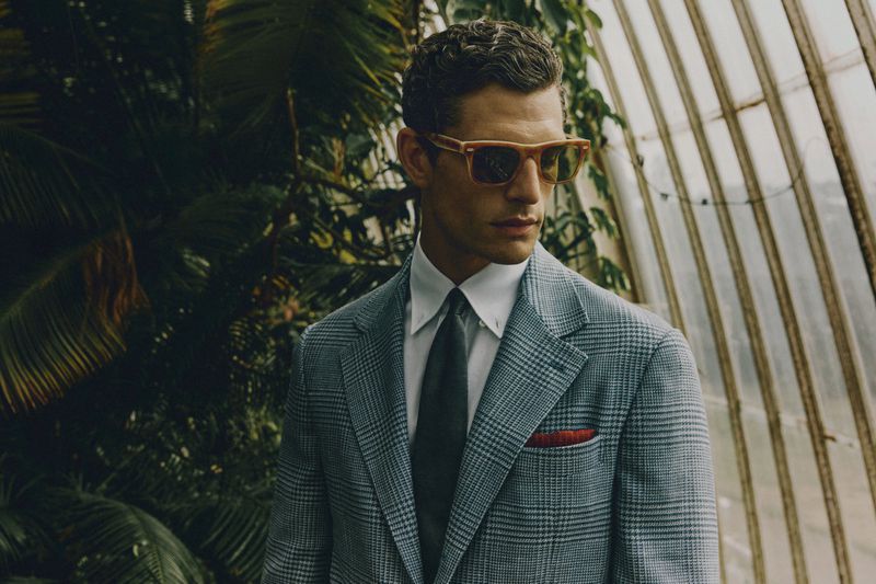 Man in a grey plaid suit and sunglasses stands in a greenhouse with tropical foliage. Fashion shot with natural light, and vintage styling.