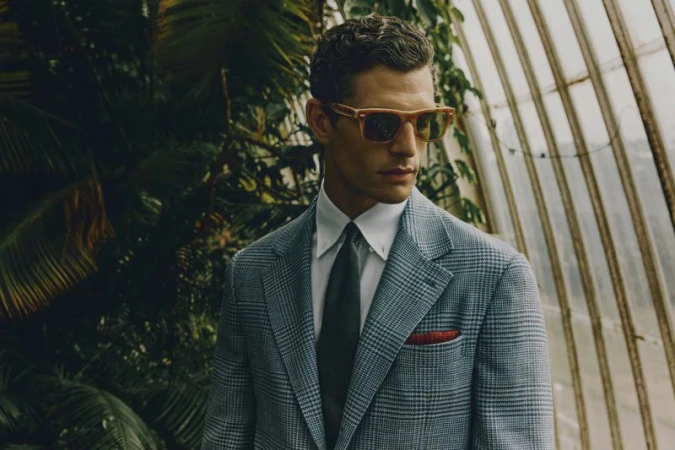 Man in a grey plaid suit and sunglasses stands in a greenhouse with tropical foliage. Fashion shot with natural light, and vintage styling.