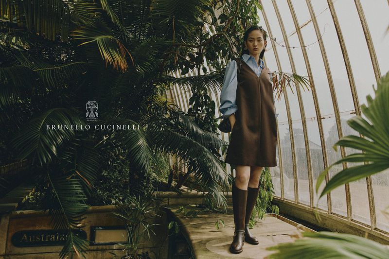 Asian woman in a brown dress and boots poses in a greenhouse surrounded by tropical plants and a glass and metal structure.