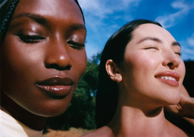 Two women pose outdoors with eyes closed, enjoying the sun's warmth on their faces in a close-up shot against a bright blue sky.