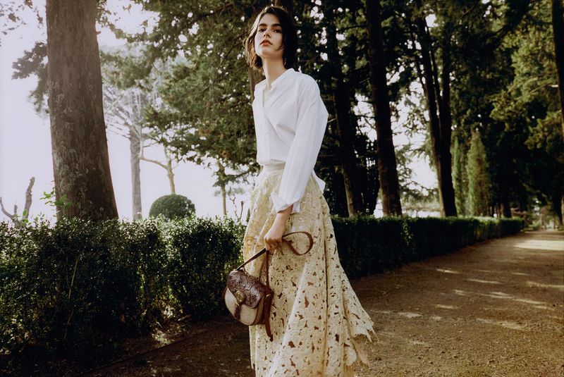Adult female model in white shirt and beige skirt holding a handbag, posing on a pathway in a park setting with tall trees.
