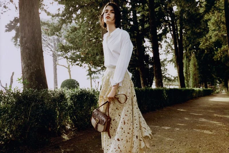 Adult female model in white shirt and beige skirt holding a handbag, posing on a pathway in a park setting with tall trees.