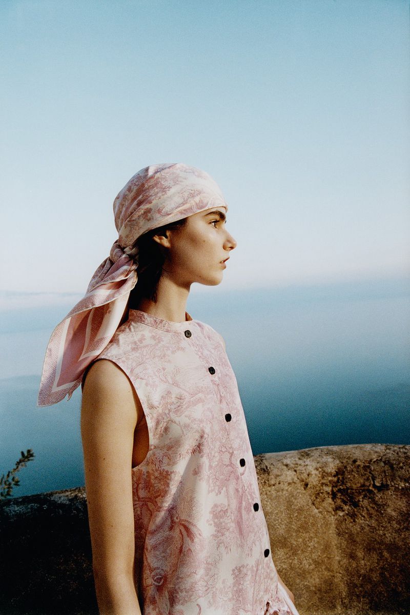Profile shot of a fair-skinned woman in a patterned headscarf and matching sleeveless dress, posing outdoors on a sunny day with a sea view.