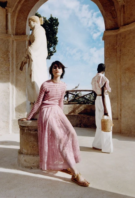 Two women in a courtyard with a statue under an arch. One woman sits in pink; the other stands in white with a wicker purse.
