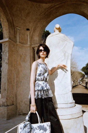 A woman in a floral top and black pants poses next to a statue in front of a building with an arched doorway on a sunny day.