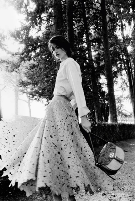 Black and white fashion shot of woman in a full skirt holding a purse outdoors in a grove of trees with sunlight shining through.