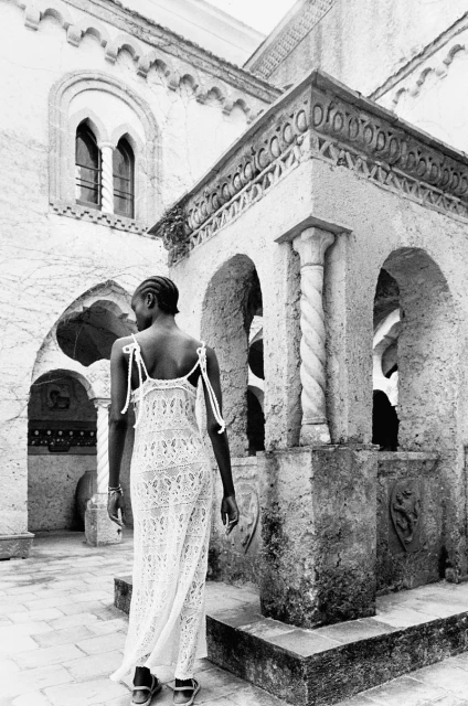Adult woman in a long lace dress walks on a stone tile patio. Building with arches and columns in the background in black and white.