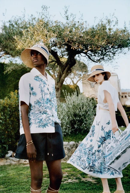 Two models pose in matching straw hats, white tops and graphic print skirt and shorts in lush outdoor setting. Fashion advertising.