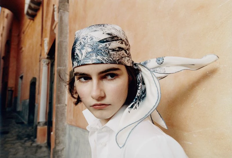 Portrait of a young woman wearing a patterned headscarf, a white blouse, with an alleyway background. Fashion and portrait photography.