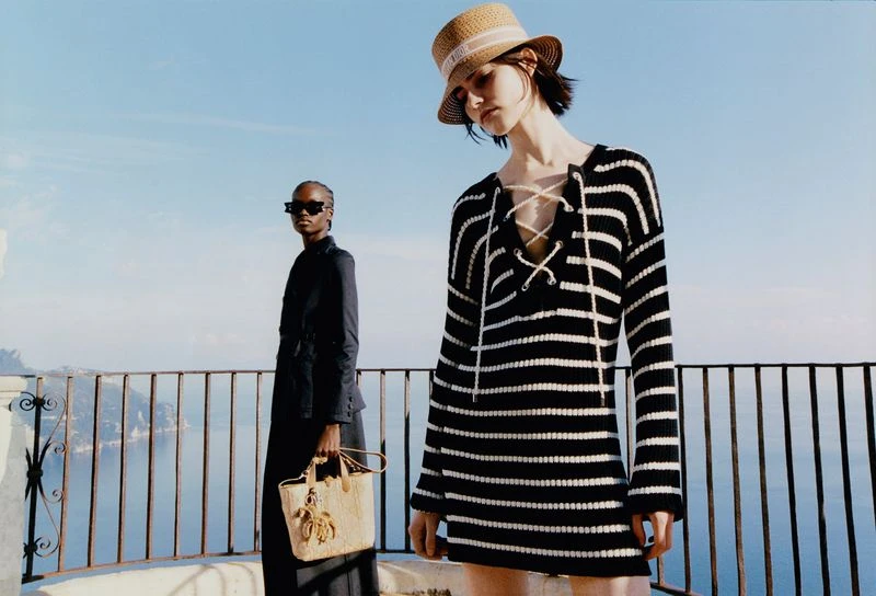 Two fashion models pose on a balcony overlooking a seascape. One wears a striped sweater dress and straw hat; the other wears sunglasses and a dark suit.
