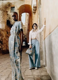 Two female models posing in a historic street wearing patterned blue silk dress and denim trousers with a handbag for Dior Riviera.