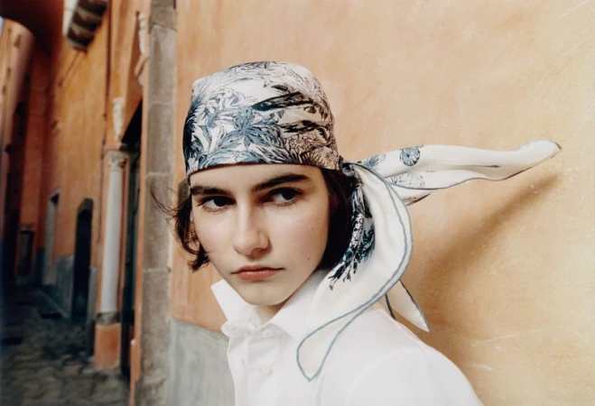 Model wearing a printed silk headscarf tied around her head and a white shirt against a warm terracotta wall.