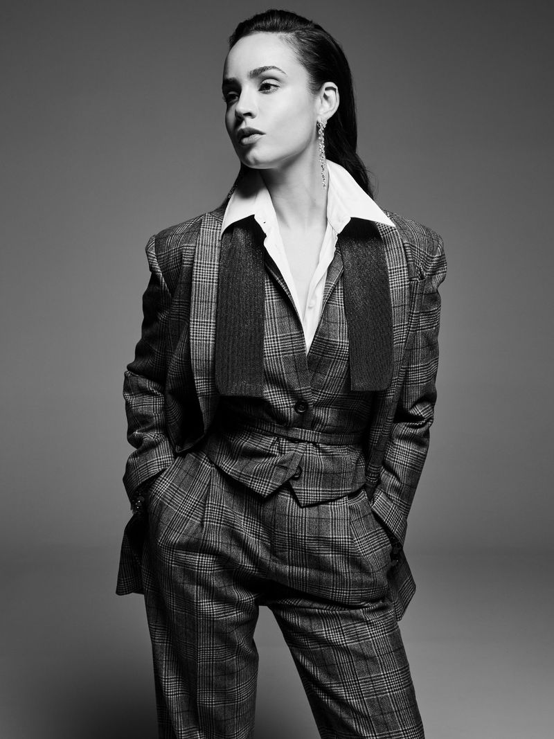 Black and white portrait of a young woman in a plaid suit with hands in her pockets, set against a grey background in a studio.