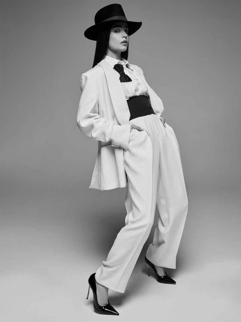 Elegant woman in wide-leg pants and a blazer poses in a photo studio wearing a black fedora, black belt, and high heels.