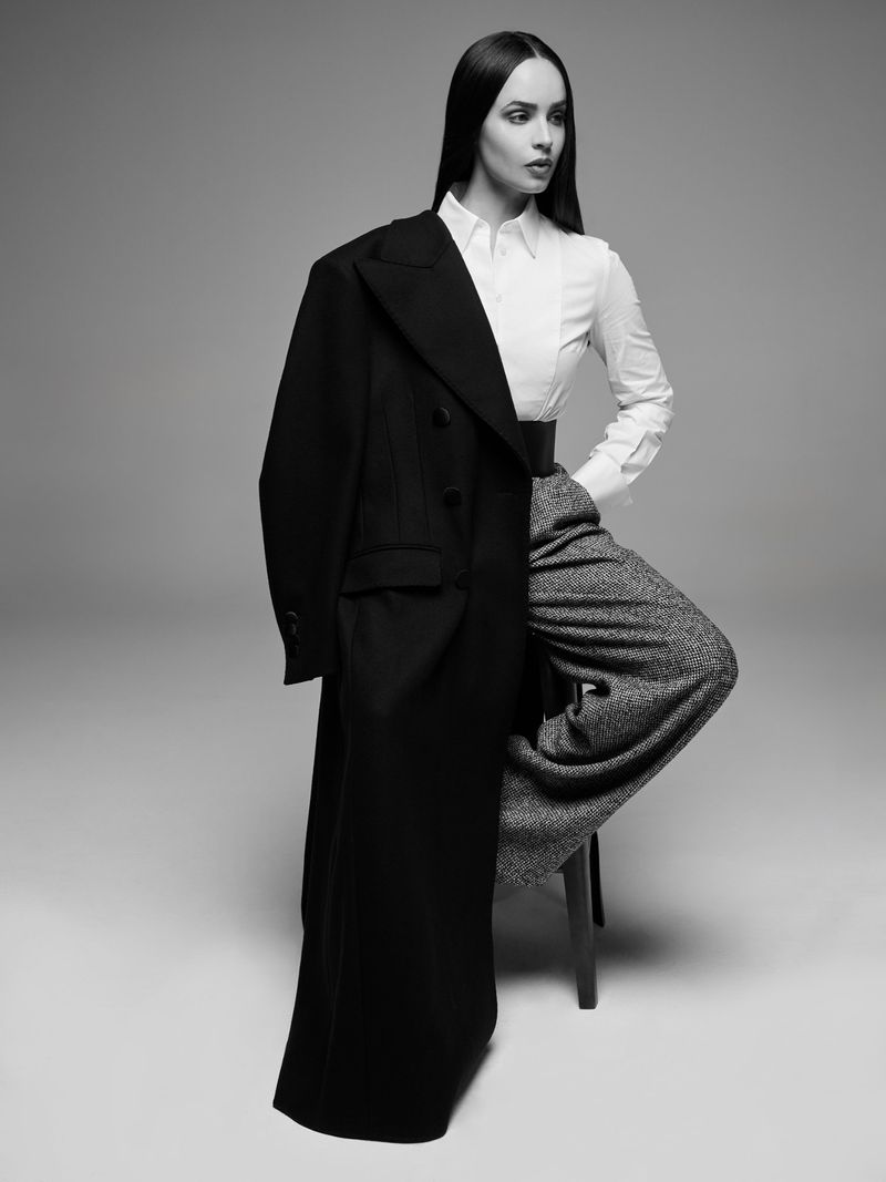 Monochrome studio shot of a woman in a white blouse and wide-legged trousers, with a black overcoat draped over her shoulder.