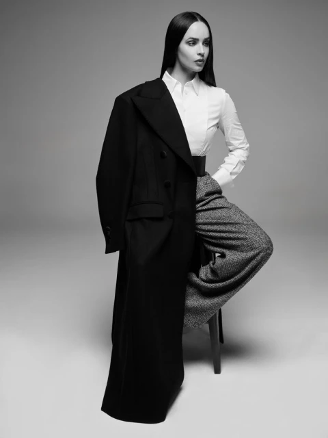 Monochrome studio shot of a woman in a white blouse and wide-legged trousers, with a black overcoat draped over her shoulder.