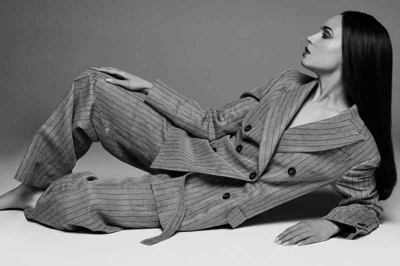 Woman in a grey striped pantsuit reclines in a photo studio. Her long, dark hair contrasts with the suit and the neutral backdrop.