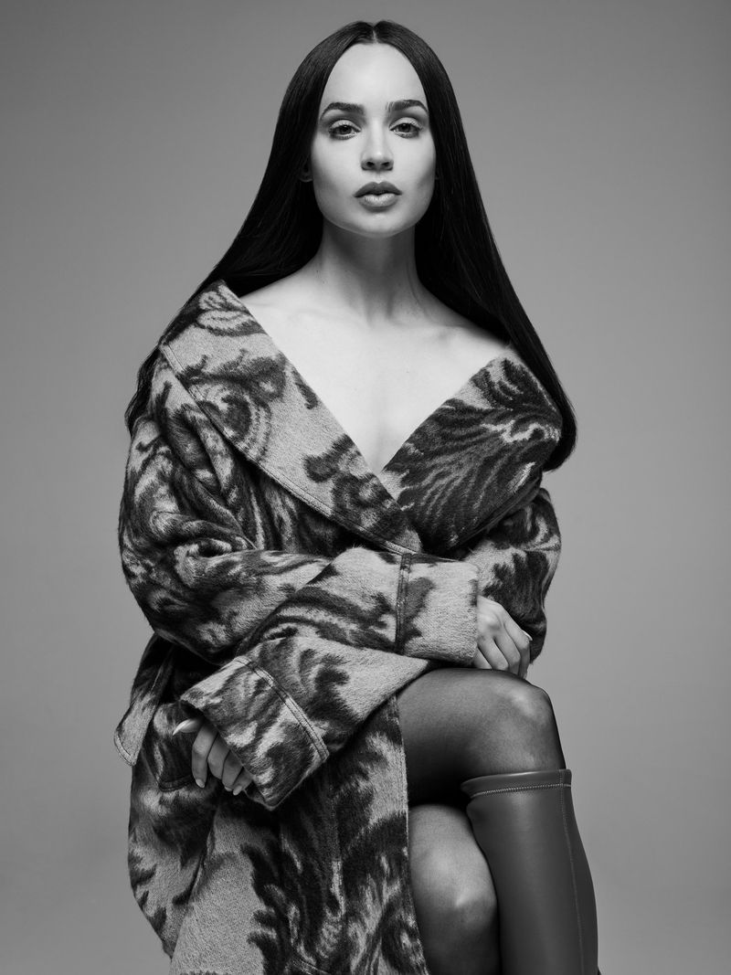 Black and white portrait of Sofia Carson wearing a patterned off-the-shoulder coat and boots against a studio backdrop.