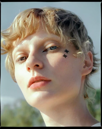 Close-up portrait of a young adult female with heterochromia, fair skin, blonde hair, and face tattoos, looking directly at the camera.