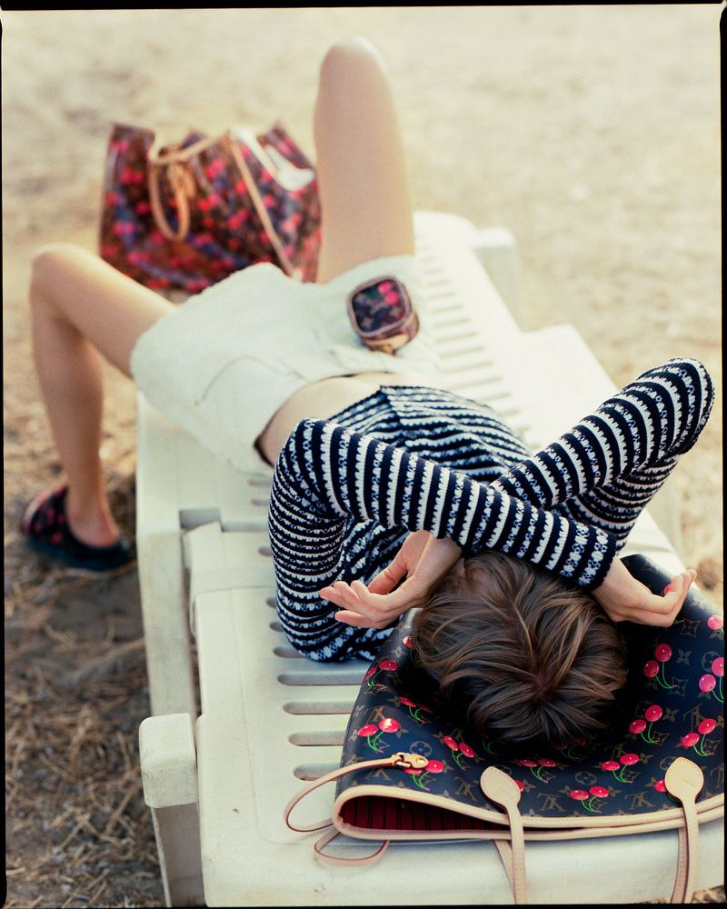 Fashion shot of a woman lying on a lounge chair at the beach with designer handbags. The model is wearing white shorts and striped top.