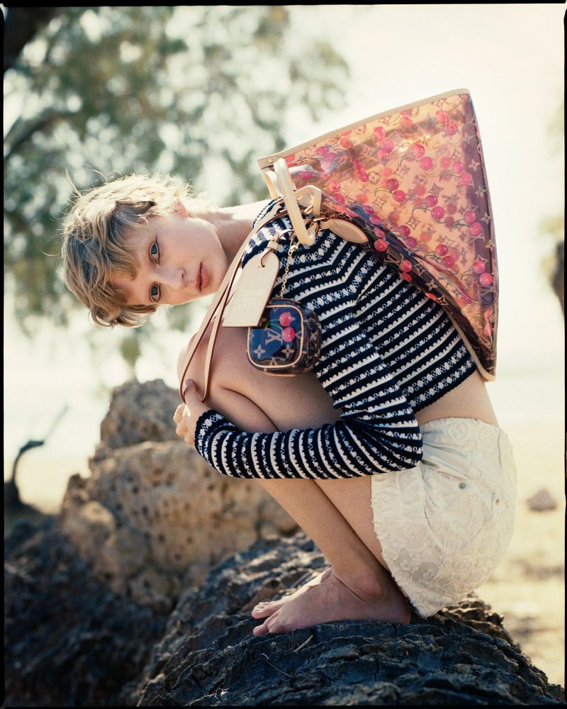 Barefoot woman crouches on a rock with transparent Louis Vuitton handbag and small purse in an outdoor, sunny, seaside environment.